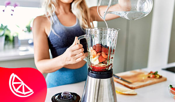 Une dame en tenue de sport ajoute de leau dans un blender dont le bol contient des morceaux de banane, de fraises, de framboises et de myrtilles.