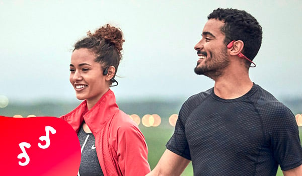 Een man en een vrouw in sportieve kledij wandelen glimlachend. Ze dragen beiden sportoortjes, zwarte voor haar en roze voor hem.