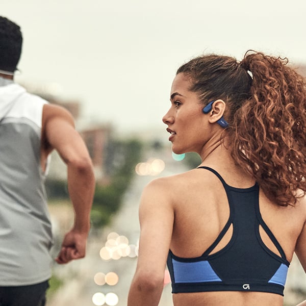 Deux personnes en tenue de sport font leur jogging prs d'une route frquente. Au premier plan, on voit une femme portant des couteurs Shokz OpenRun bleu fonc qui regarde lgrement de ct.