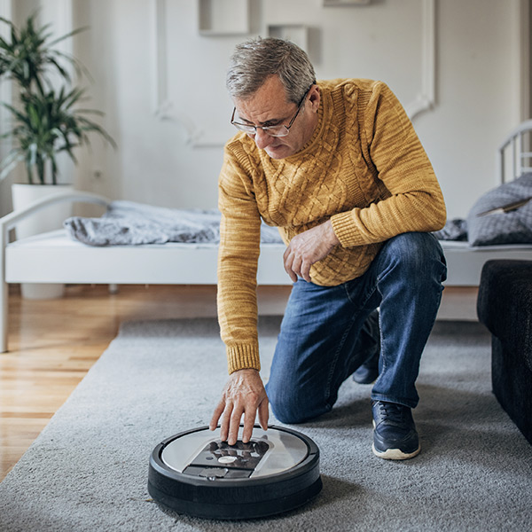 Un homme portant des lunettes, un pull jaune et un jean fait fonctionner son robot aspirateur